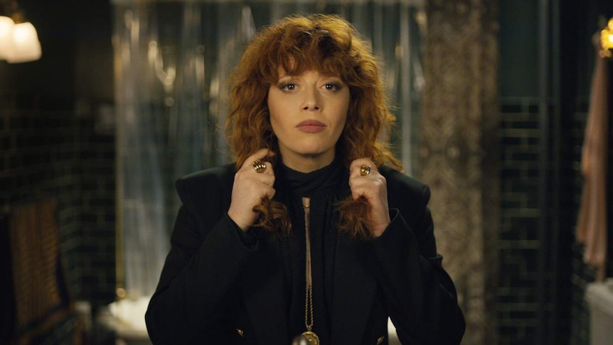 Woman with curly red hair standing facing camera as if looking at herself in mirror, holding her hair with each hand.