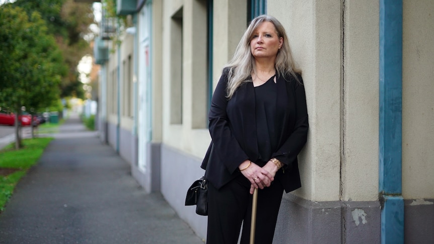 Donna leans against the wall of a building holding a cane in her hands.