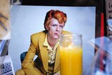 A photo of David Bowie, squatting and wearing a yellow suit, sits among flowers at a memorial.