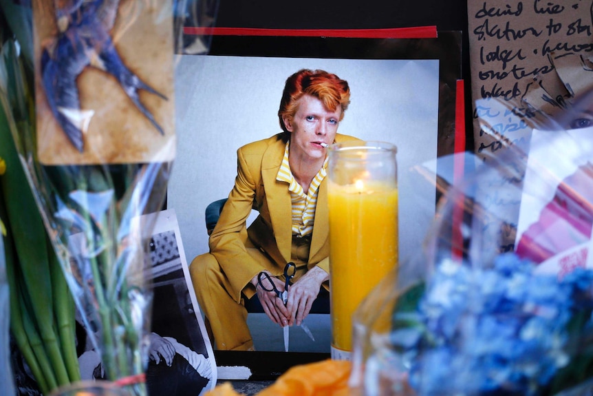 A photo of David Bowie, squatting and wearing a yellow suit, sits among flowers at a memorial.