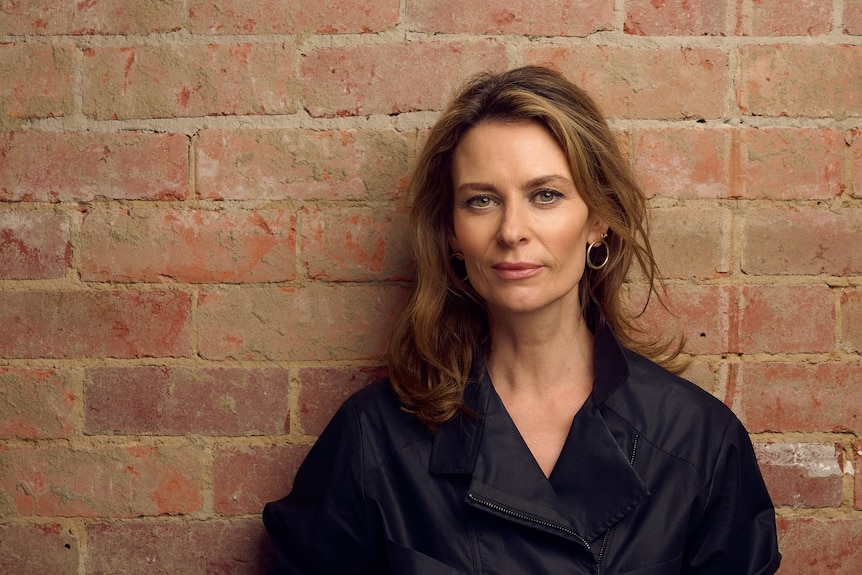 The actor Kat Stewart, a woman in her early 50s with long brunette hair, standing in front of a brick wall