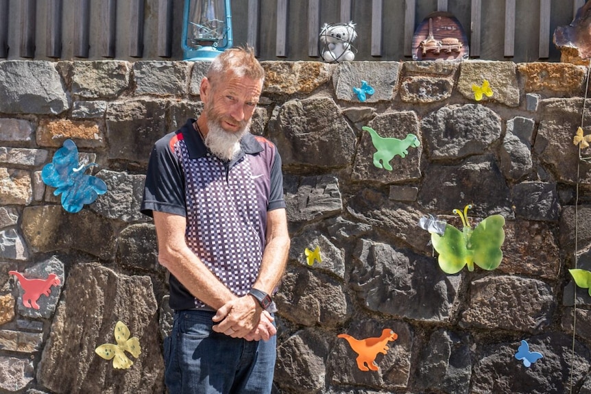 A man with a grey beard stands next to his courtyard wall that is covered in artistic butterflies.