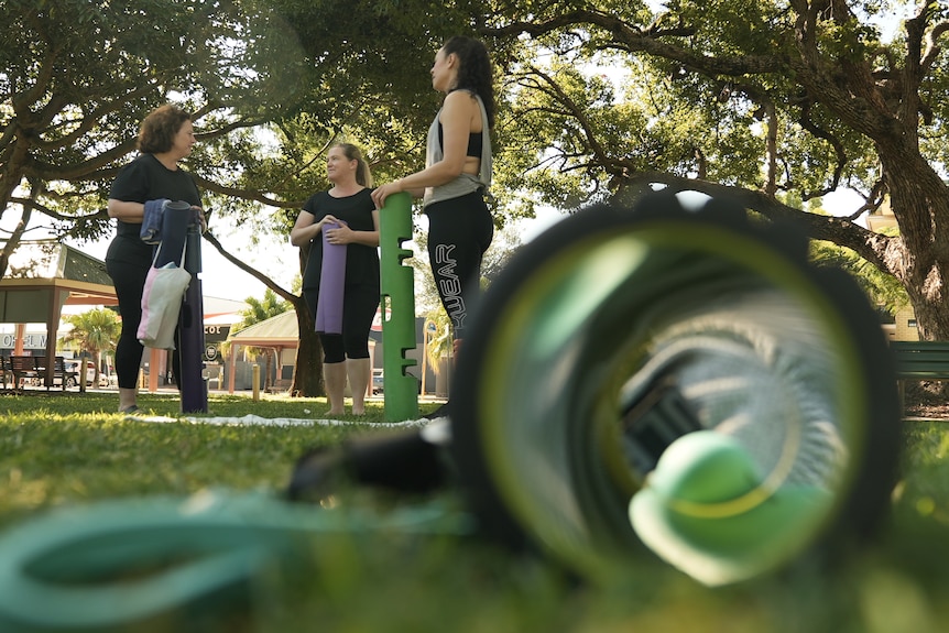 Three women stand together in a park with exercise equipment nearby.