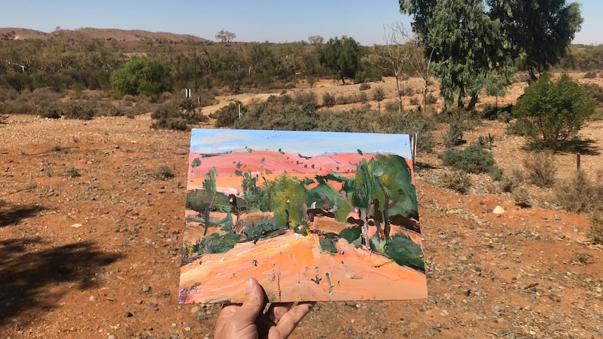 A painting is held up in front of the landscape, the area is dry with orange sand and some shrubs