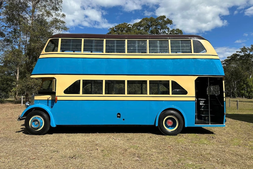 The blue and cream double decker bus is parked side on in a large grassed area.