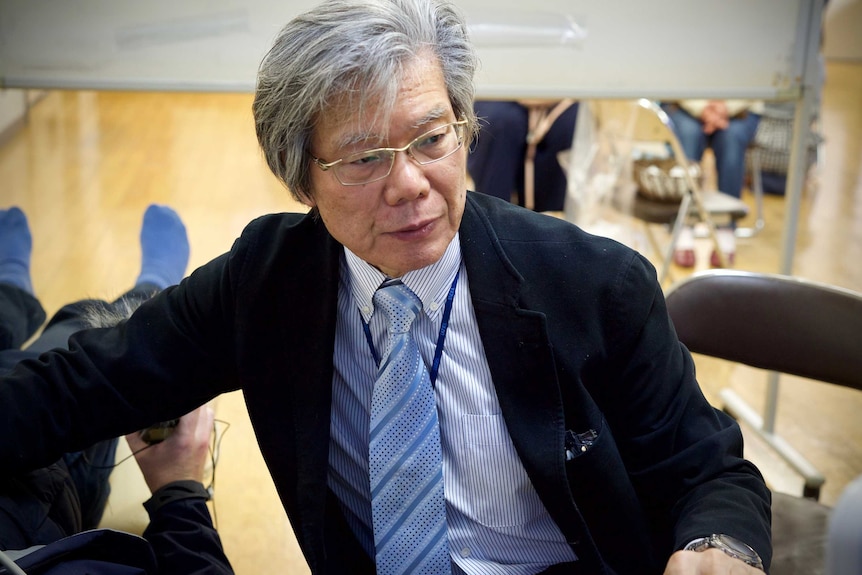 A Japanese man in a suit and gold glasses looks at a monitor