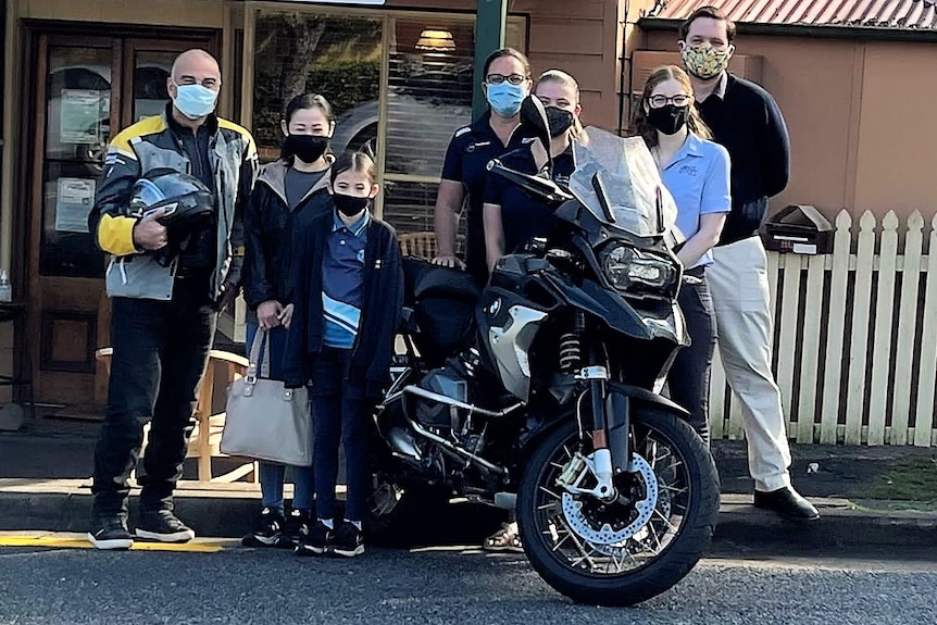 A group of people, wearing face masks, standing outside around a motorbike.