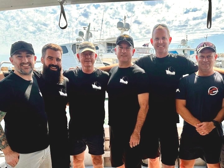 Six men in black tshirts stand smiling in a row on a boat.
