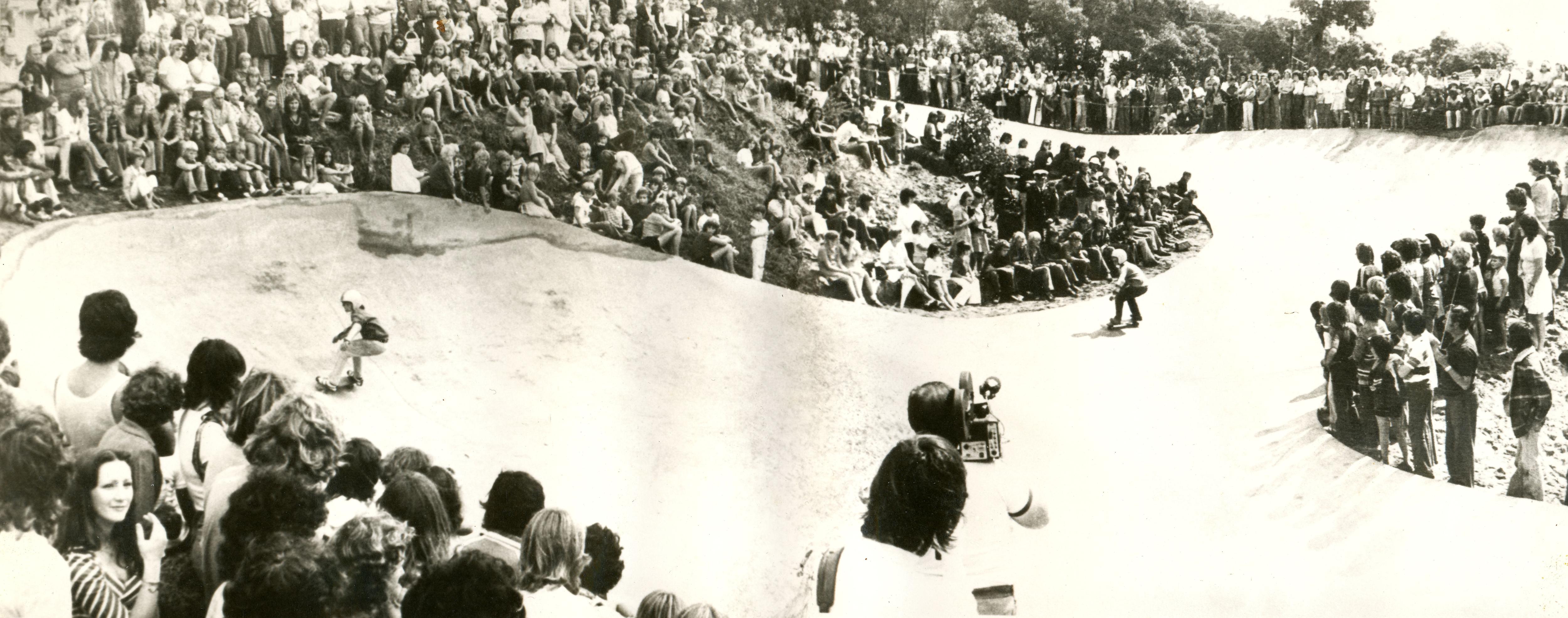 Una fotografía en blanco y negro de multitudes observando a la gente patinando en un parque de patinaje. 