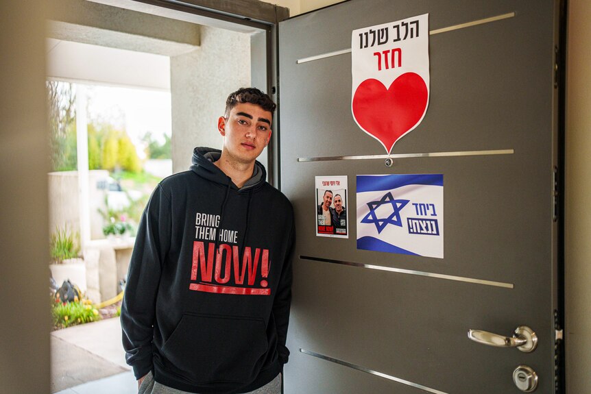 Ofir Engle wears a jumper saying Bring Them Home Now while leaning against a door.