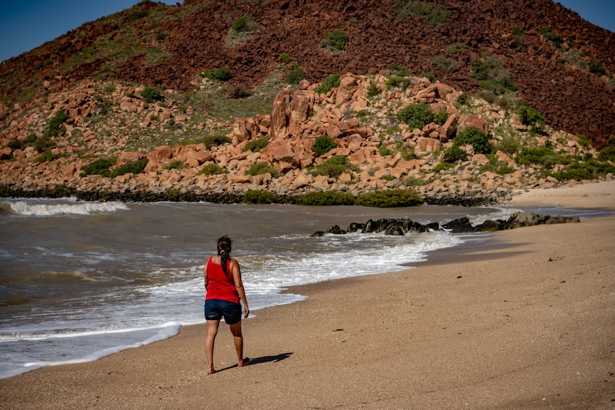 Raelene Cooper camina por la playa, con rocas al fondo.