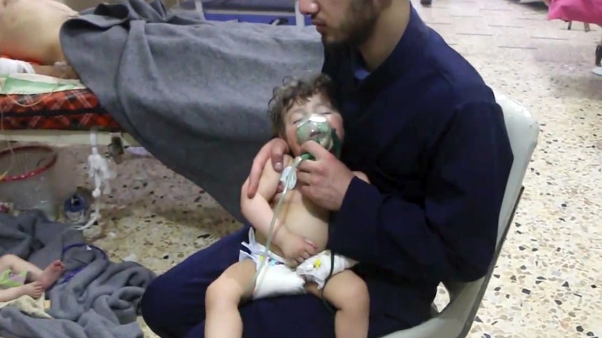 A medical worker gives a toddler oxygen. Someone is laying in a bed beside him. There are patients on the floor, one is a child.