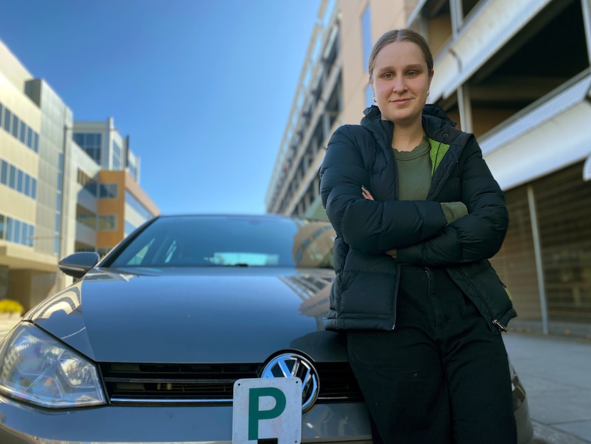 A woman leans on the bonnet of a car, with a green P-plate on it.