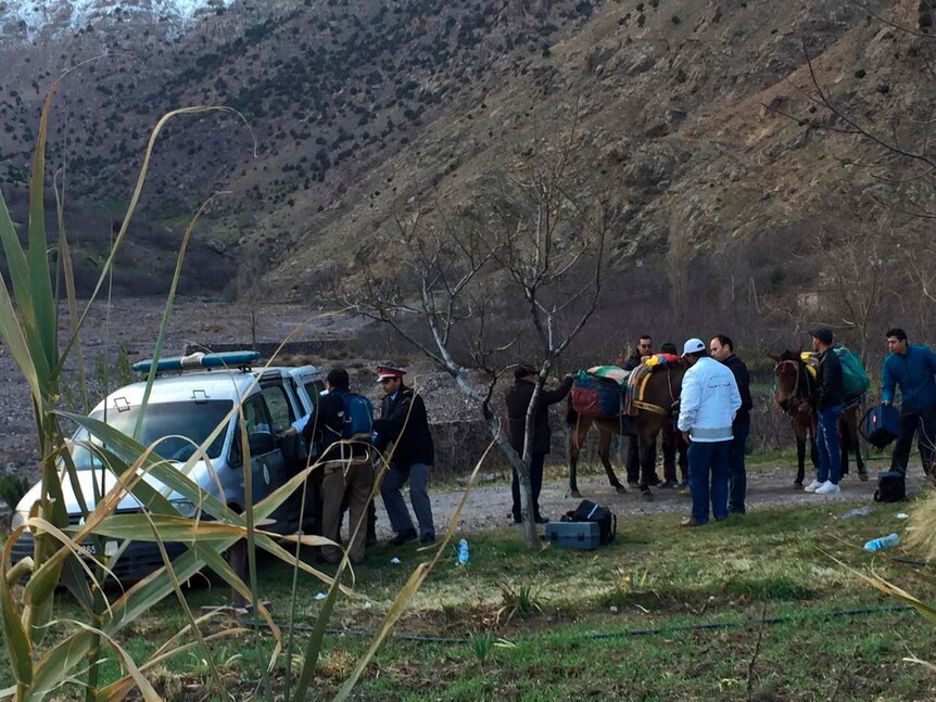 A group of people stand around a police car and two donkeys at the foot of a mountain.