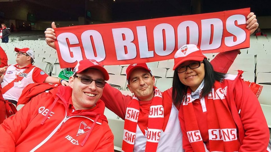 Three people dressed in red and white Swans themed clothing, with one holding up a sign that says "Go Bloods"