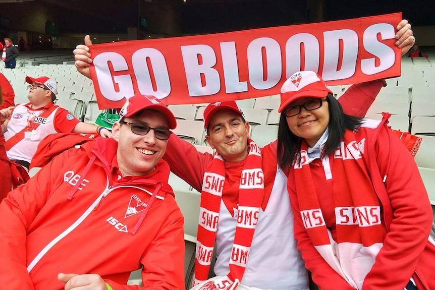 Three people dressed in red and white Swans themed clothing, with one holding up a sign that says "Go Bloods"