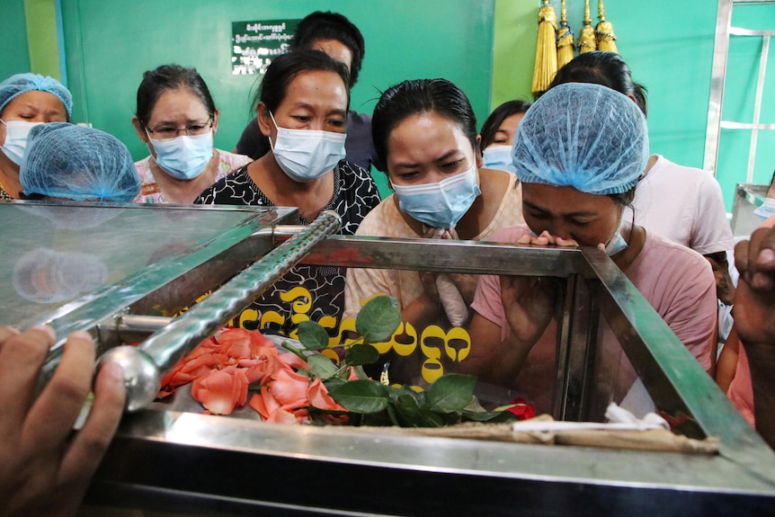 A group of female mourners wearing blue facemasks surround an open coffin