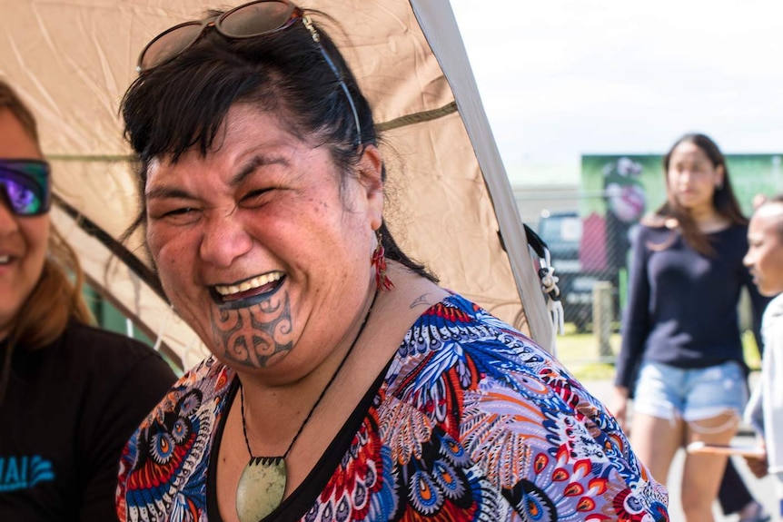 A New Zealand woman with a chin tattoo laughs and holds a plastic cup of water.