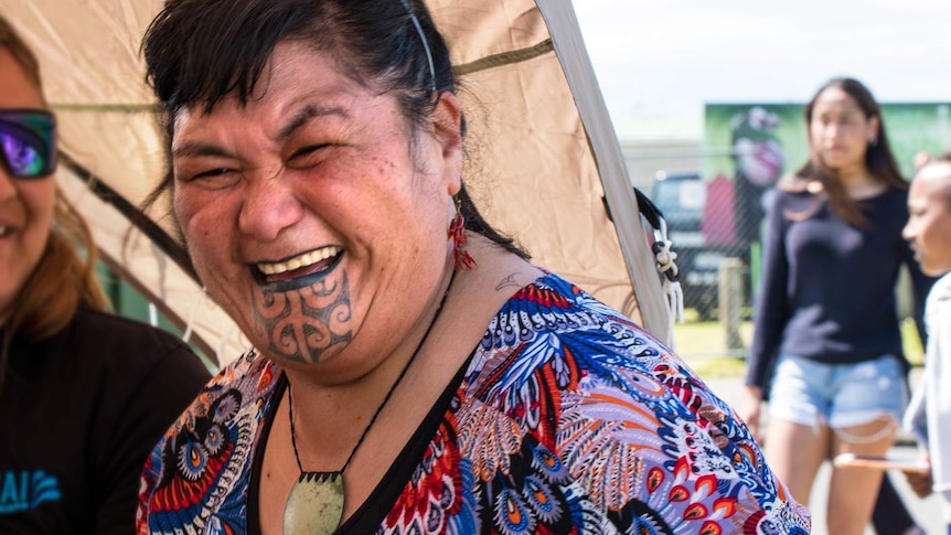 A New Zealand woman with a chin tattoo laughs and holds a plastic cup of water.