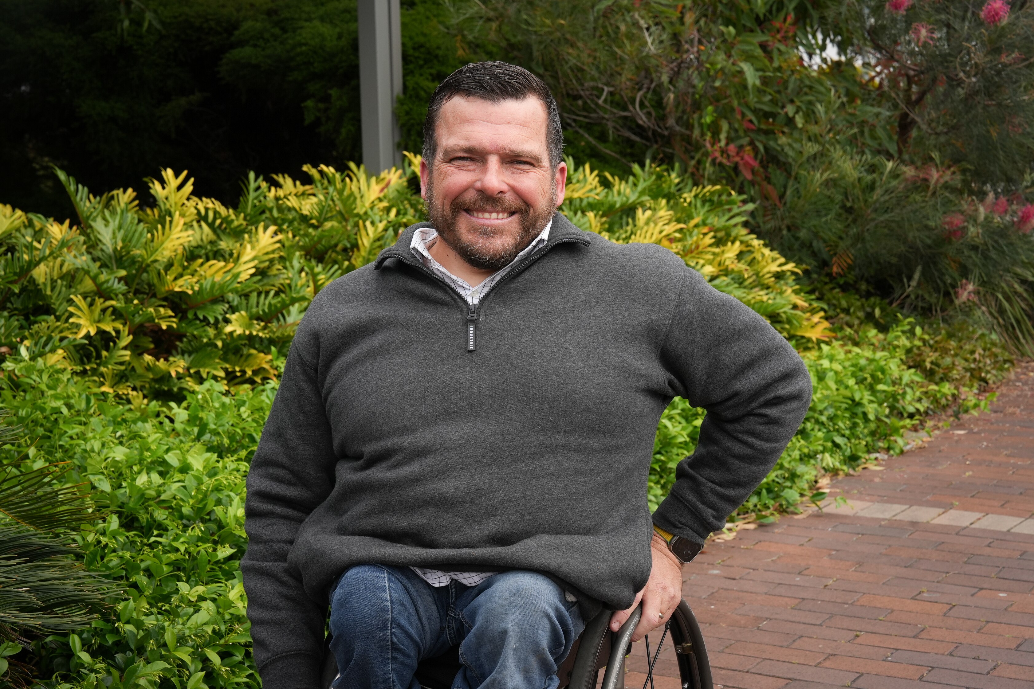 A white man with short hair and a beard sitting in a wheelchair on a footpath