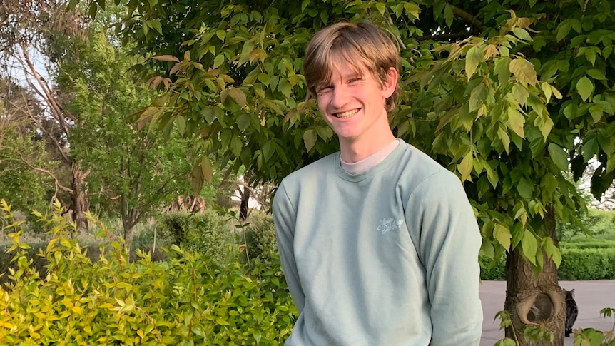 A young man stands in front of a tree smiling