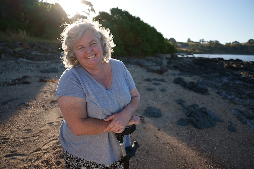 Woman in a grey shirt standing on a beach and leaning on a crutch.