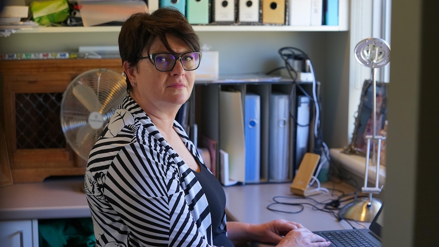 A middle-aged woman with short hair and glasses looks into camera with a serious expression.