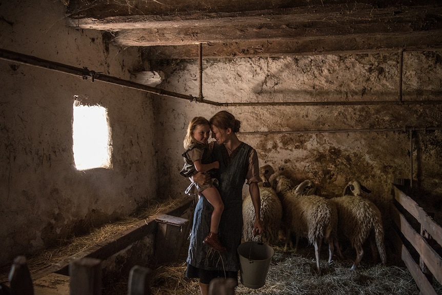 In front of sheep, woman stands holding bucket in one hand and rests young daughter on other hip inside dimly lit concrete shed.