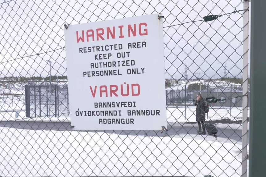 A warning sign on a barbed wire fence of a prison with snow on the ground and a woman wheeling a suitcase in the background