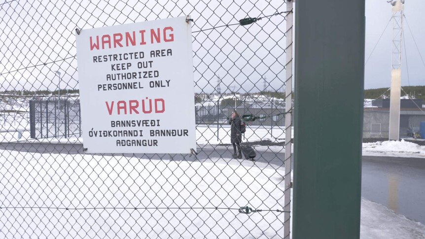 A warning sign on a barbed wire fence of a prison with snow on the ground and a woman wheeling a suitcase in the background
