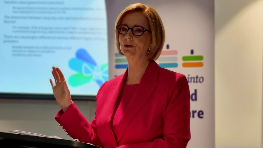 Julia Gillard stands at a podium speaking with a powerpoint display behind her