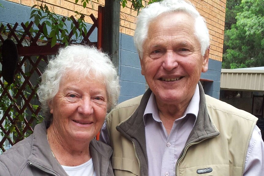 An elderly couple stands outside and smiles at the camera