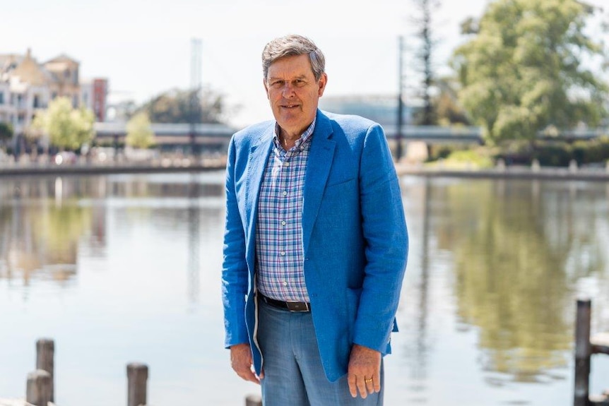 A medium shot of Tim wearing a blue suit in front of a water inlet, with trees and a bridge out of focus in the background.