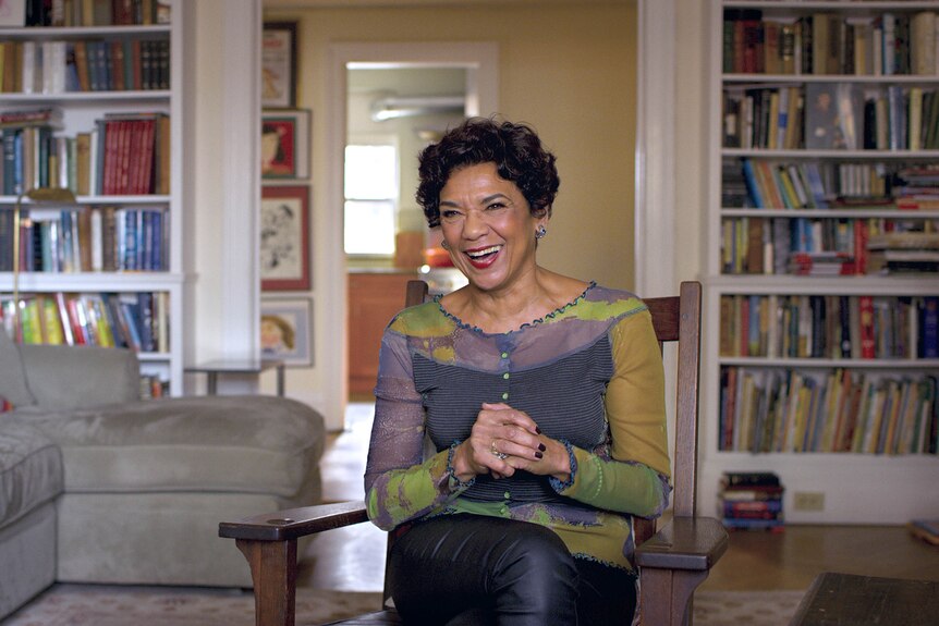 Older black woman with short dark hair and beaming smile, wears purple and green shirt and sits in home with bookshelves.