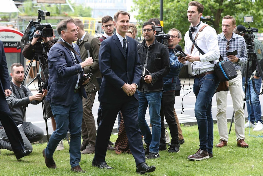 Jeremy Hunt in a suit walks across grass surrounded by press with microphones and cameras.
