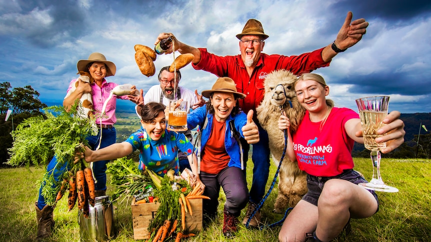 Six people and a alpaca on a hill top holding up produce including carrots, beer, wine and bread. 