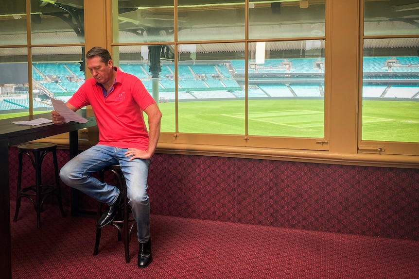 man in dark pink polo shirt sits on a stool in an empty carpeted room overlooking a cricket field