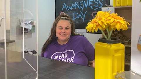 A woman in a purple T-shirt that reads "#End overdose" sits behind a counter, smiling