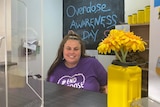 A woman in a purple T-shirt that reads "#End overdose" sits behind a counter, smiling