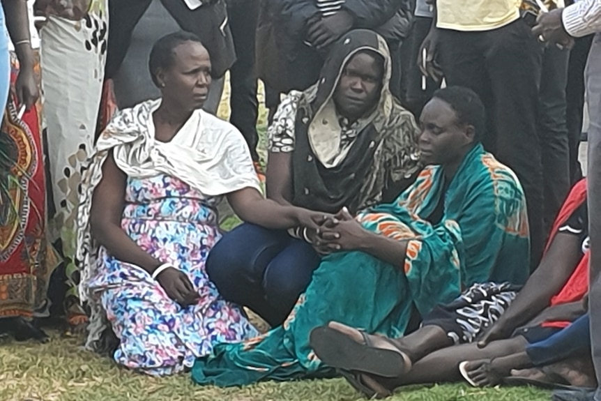 Aguer Akec's mother wears a green and orange shawl and sits on the ground with two women beside her holding her hands.