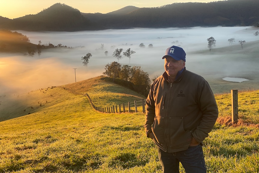 A man, wearing a blue cap, stands on his farm with fog in the valley behind him.
