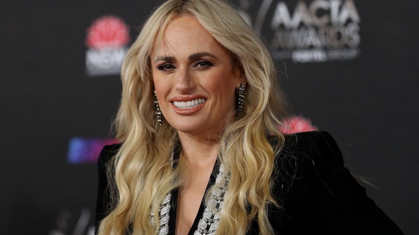 Woman with long white blonde hair wears flattering makeup and a black should-padded dress on a red carpet.