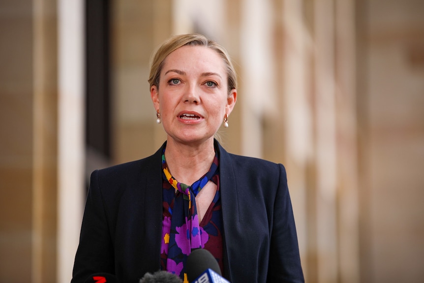 A woman with short tied back hair, a coloured scarf and a jacket speaking