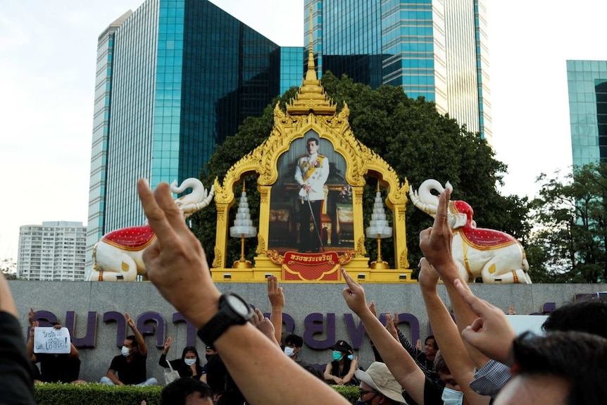 People raise a two-finger salute pointing in the direction of an ornate portrait of King Vajiralongkorn in front of skyscrapers.