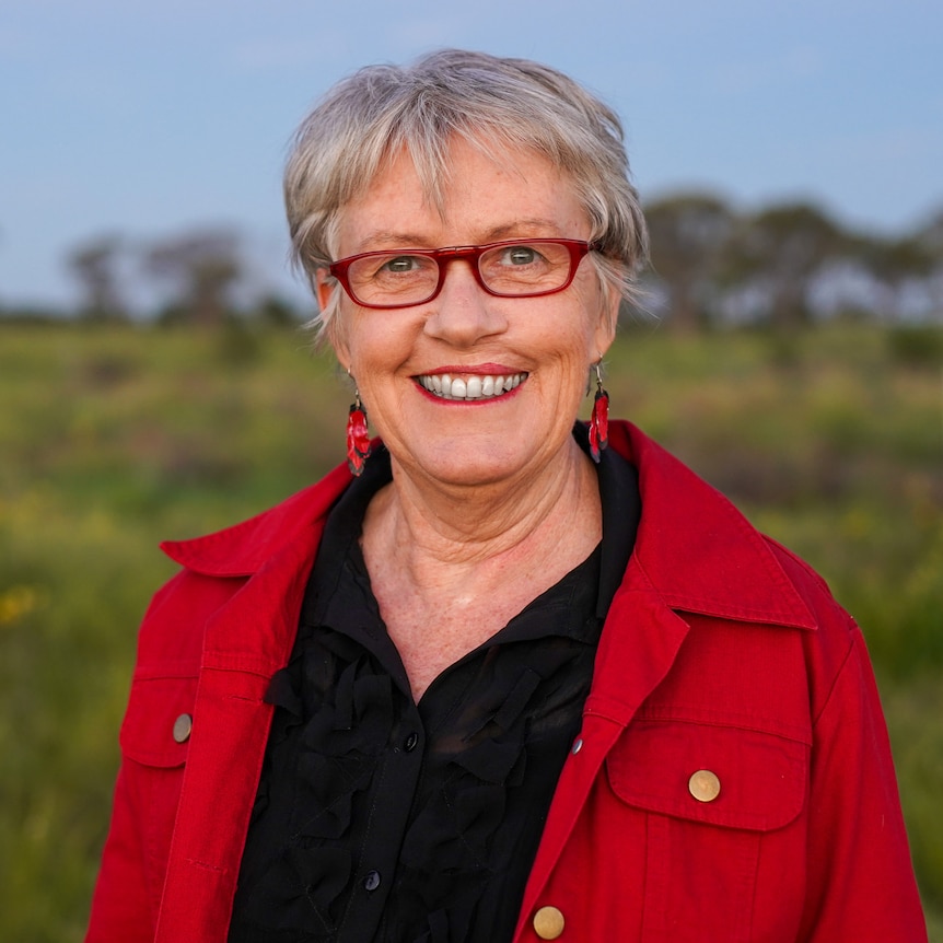 A smiling women with grey hair wearing red glasses, earrings and shirt, with a treed landscape in the background.