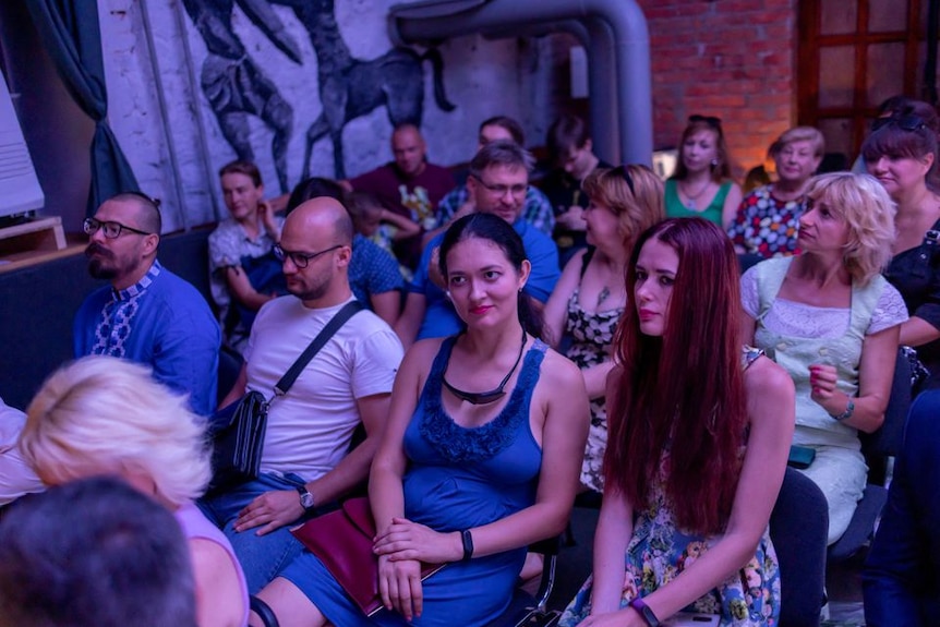 A dozen audience members sit at an underground shelter turned to an underground temporary stage and wait. 