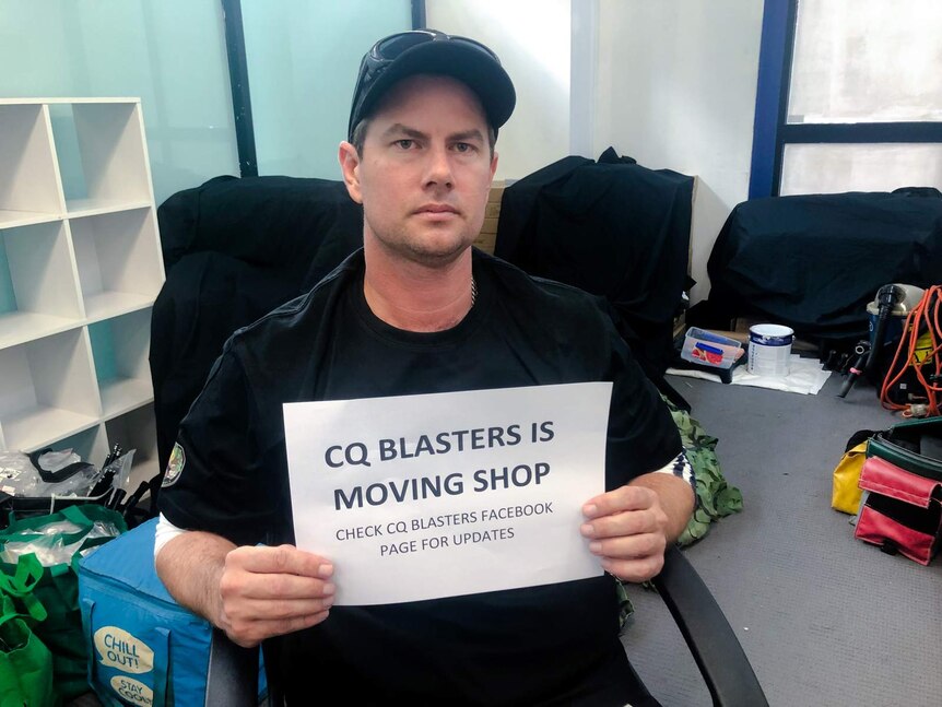 A man holds a sign 'CQ Blasters is moving shop', looks unhappy.