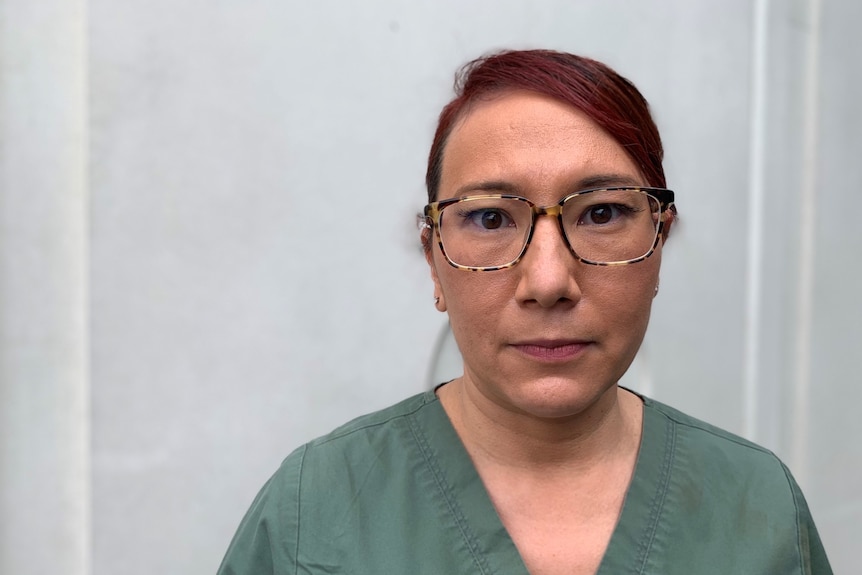 Headshot of woman with glasses and hair pulled back, wearing olive green scrubs in front of white background.