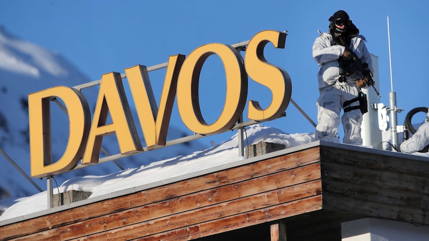 A man in a white suit holds a gun next to a sign that reads "Davos"