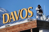A man in a white suit holds a gun next to a sign that reads "Davos"